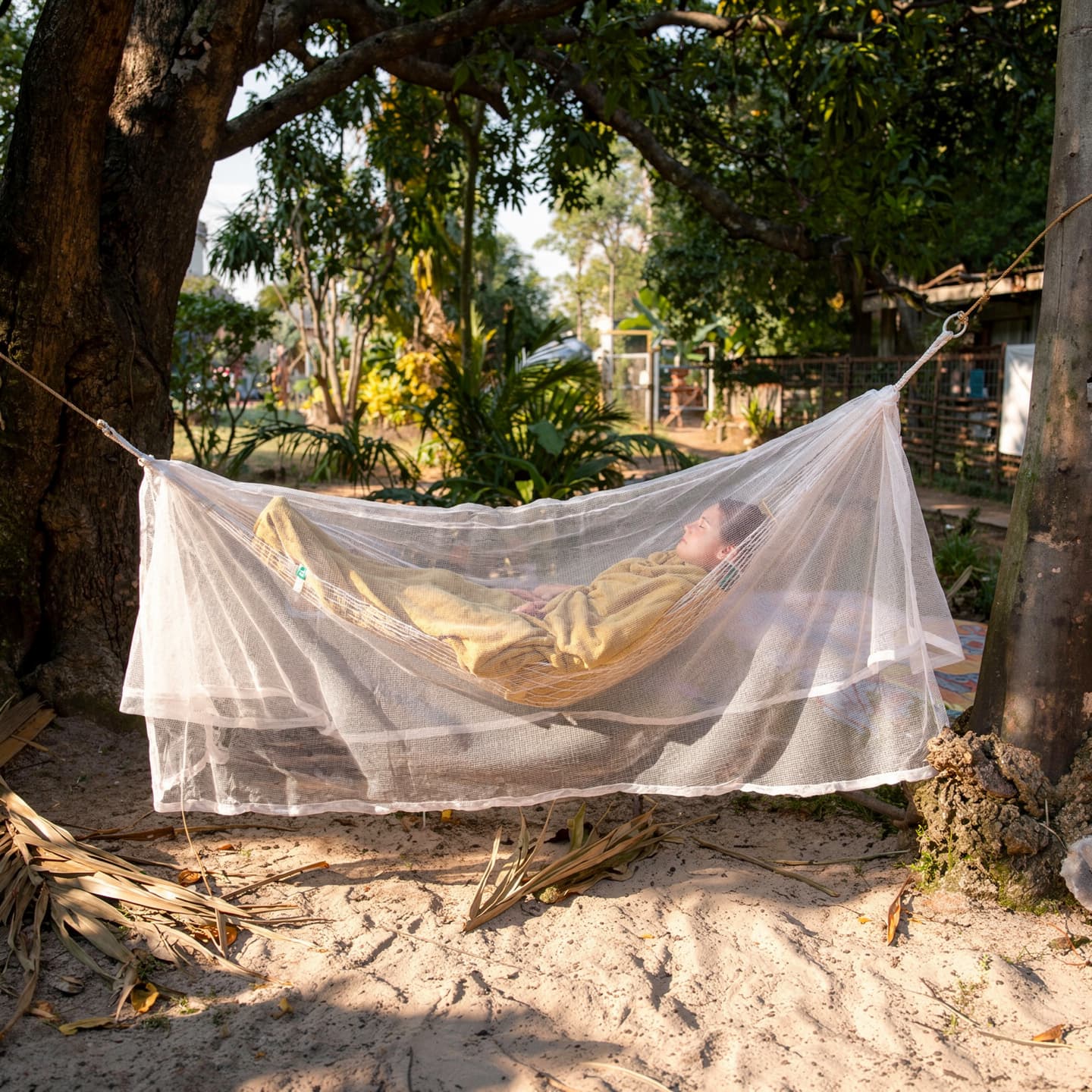 A person resting in a hammock wrapped in the soft off-white Mosticare Amaca mosquito net, in a sunlit tropical garden.
