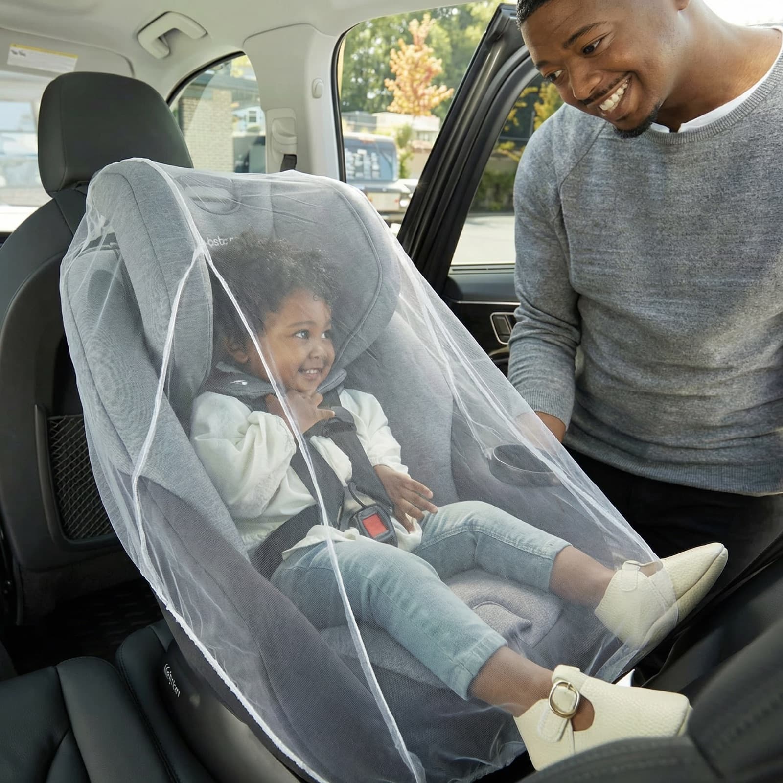 A smiling father beside a curly-haired toddler in a car seat shielded by the Mosticare Bambino Carello stroller mosquito net.