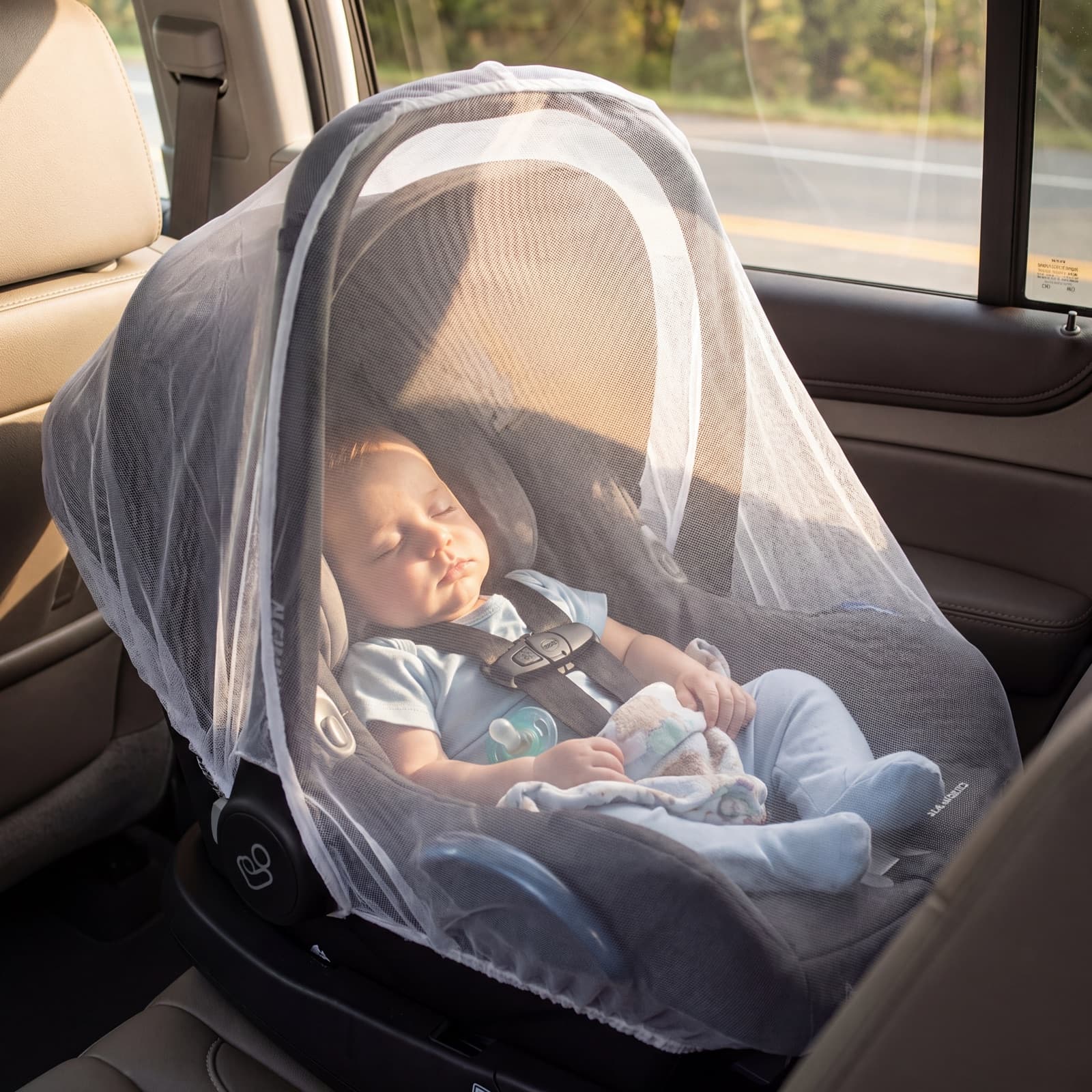 A baby asleep in an infant car-seat carrier draped with the Mosticare Bambino Carello mosquito net in warm afternoon light.