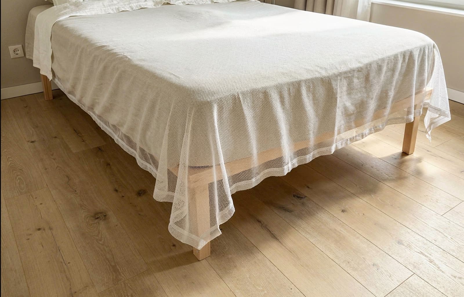 The Mosticare Materasso mosquito-net envelope draped over a mattress in a calm, sunlit modern bedroom.
