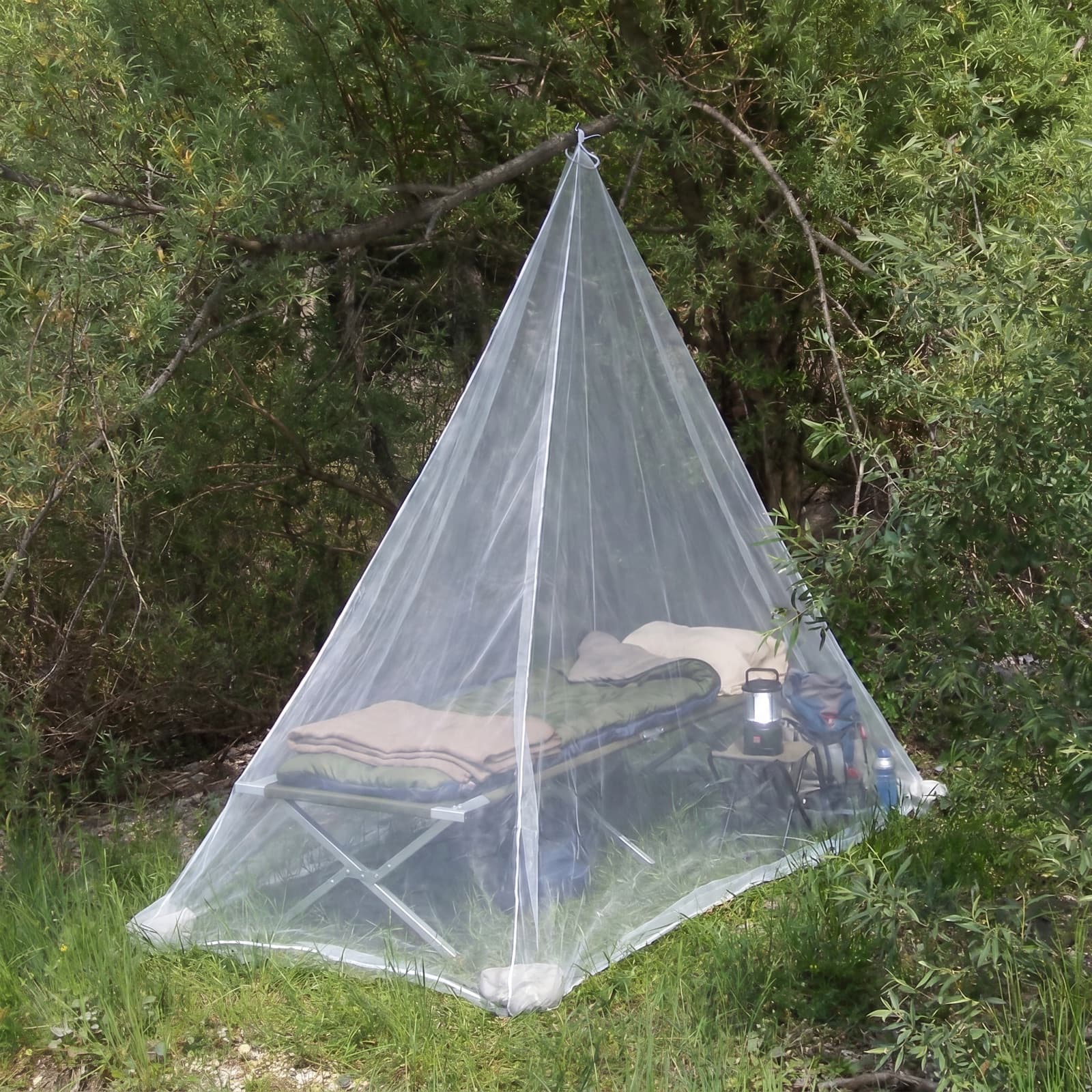 The Mosticare Traccia travel mosquito net pitched over a camp bed and lantern at a woodland campsite.