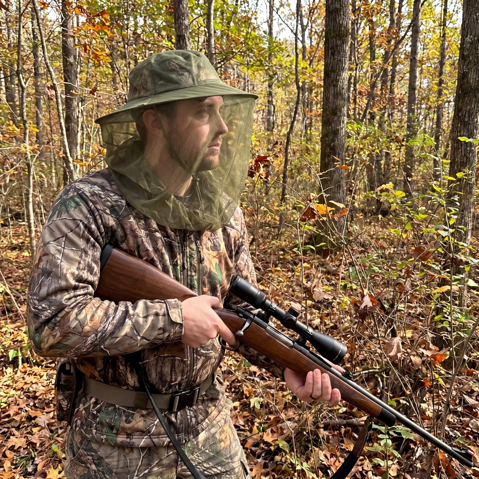 A person in camouflage wearing the green Mosticare Volto head net over a wide-brim hat in autumn woodland.